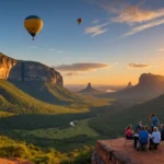 Vista aérea de um grupo de pessoas sobre uma pedra no alto da serra, com vale entre morros e montanha ao fundo sob o céu azul